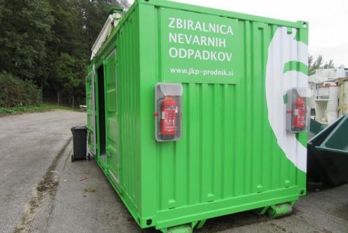 Premična zbiralnica - kontejner za nevarne odpadke, zelene barve z belim napisom in gasilnima aparatoma.