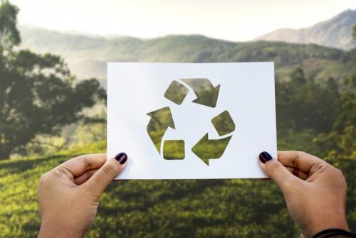 Roki držita list papirja z izrezanim znakom za recikliranje (tri puščice), skozi katerega se vidi zelena narava v ozadju.